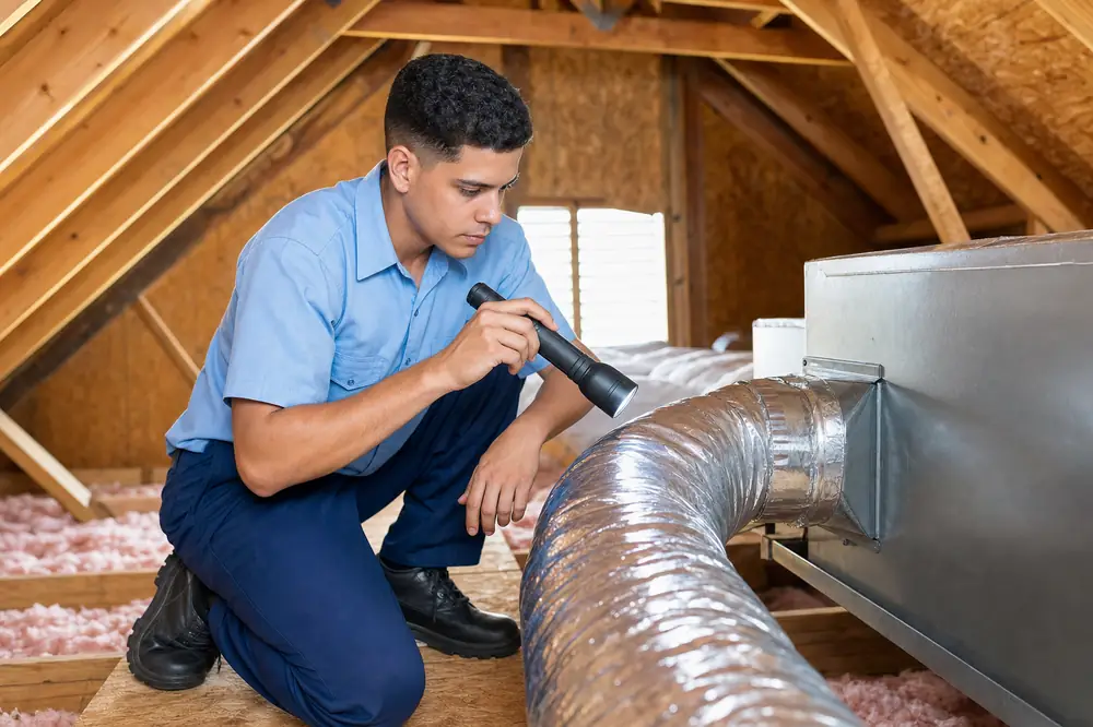 A technician inspecting a flexible duct joint in an attic with a flashlight